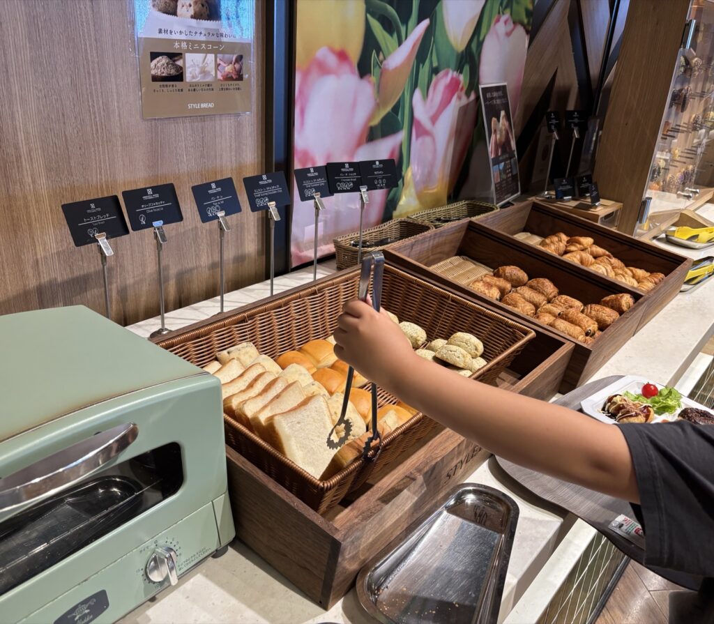 ホテルの朝食ビュッフェでスタイルブレッド(STYLE BREAD)のパンをトングで取る子どもの手。パン・オ・レやミニスコーンの名札が並ぶ